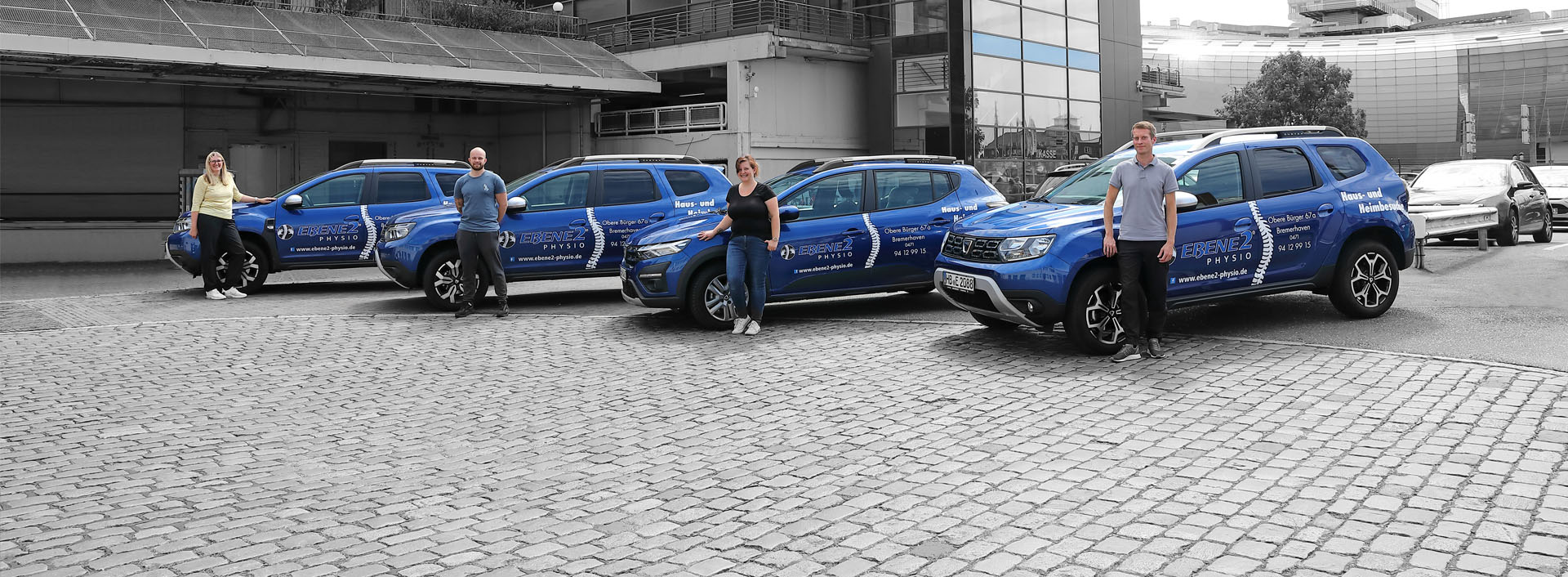 Hier sieht man vier Fahrzeuge der EBENE2 Physio vor dem Columbus Center in Bremerhaven. Die Physiotherapeuten posieren davor. Im Hintergrund sieht man das Sail City Hotel und das Klimahaus.