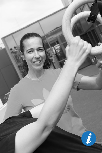 Auf dem Bild sieht man eine Frau, die in Ringen hängt. Man sieht nur ihre Unterarme. Im Hintergrund steht eine Physiotherapeutin und lächelt in die Kamera. Dabei hat sie ihre Hände auf dem Rücken der Frau.