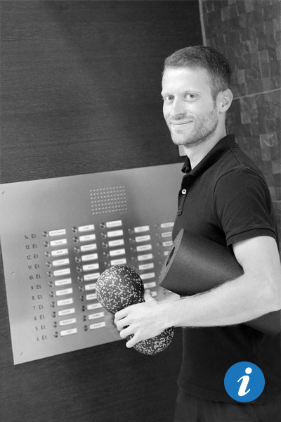 Ein junger Physiotherapeut steht an einem Klingelschild und hat in der Hand und unter dem Arm Massagerollen dabei.
