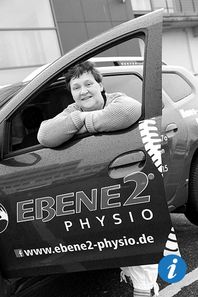 Eine Physiotherapeutin steht an der offenen Fahrzeugtür und schaut durch die heruntergedrehte Scheibe.