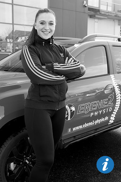 Eine hübsche Physiotherapeutin, die vor ihrem Auto für Haus- und Heimbesuche steht und in die Kamera lächelt.