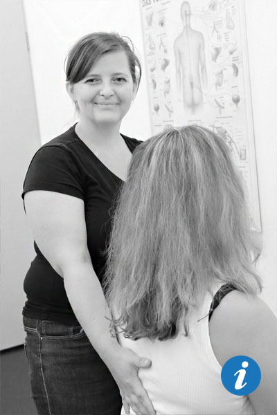 Auf dem Bild ist eine Frau zu sehen, von der nur Schulter, Hals und Kopf zu sehen sind. Sie sitzt auf einer Behandlungsliege. Im Hintergrund steht eine Physiotherapeutin und lächelt in die Kamera. Dabei hat sie ihre Hand auf dem Rücken der Frau gelegt.