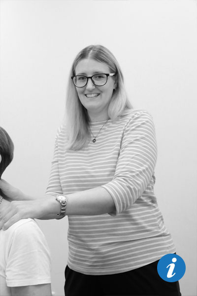 Auf dem Bild ist eine Frau zu sehen, von der nur Schulter, Hals und Kopf zu sehen sind. Sie sitzt auf einer Behandlungsliege. Im Hintergrund steht eine Physiotherapeutin und lächelt in die Kamera. Dabei hat sie ihre Hand auf die Schulter der Frau gelegt.