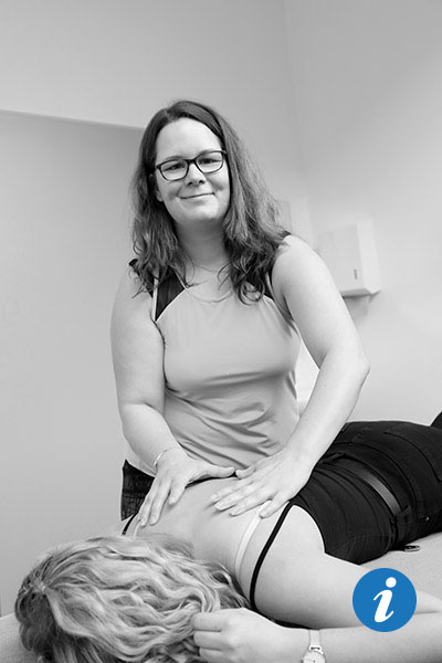 Auf dem Bild sieht man eine Frau, die auf dem Bauch auf einer Behandlungsliege liegt. Man sieht nur ihren Rücken, ihre Schultern und ihren Kopf. Im Hintergrund steht eine Physiotherapeutin und lächelt in die Kamera. Dabei hat sie ihre Hände auf den Schulterblättern der Frau.