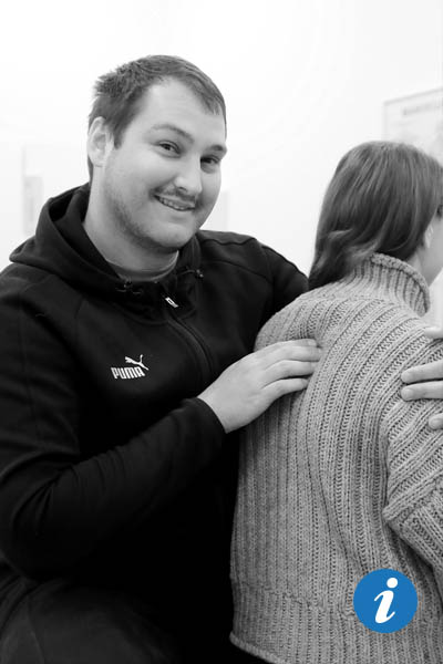 Eine Frau, von der nur Rücken, Schulter, Hals und Kopf zu sehen sind, sitzt auf einer Behandlungsliege und ein Physiotherapeut steht hinter ihr und lächelt in die Kamera, während er die Frau massiert.