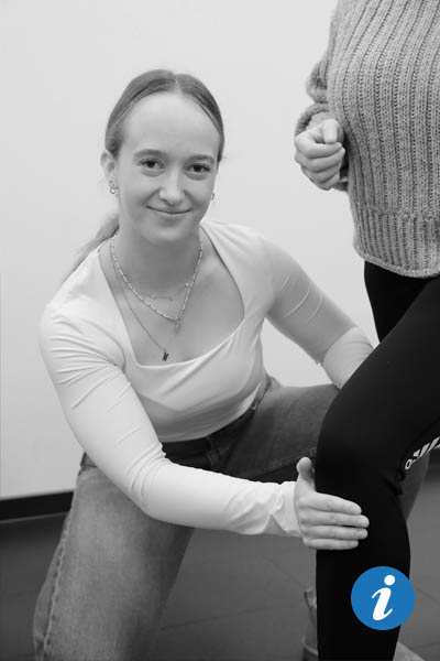 Eine Frau, von der man nur die Brust und die Beine sieht, steht vor einer Physiotherapeutin, während die Physiotherapeutin vor ihr kniet und ihr die Wade massiert.