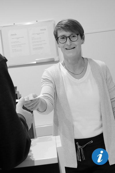 Eine Frau mit Brille und kurzem Haar steht an einem Schreibtisch und gibt einem Mann, von dem man nur die Hand sieht, einen Zettel.