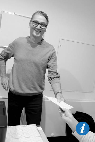 Ein Mann steht an einem Schreibtisch und gibt einer Frau, von der man nur die Hand sieht, einen Zettel.