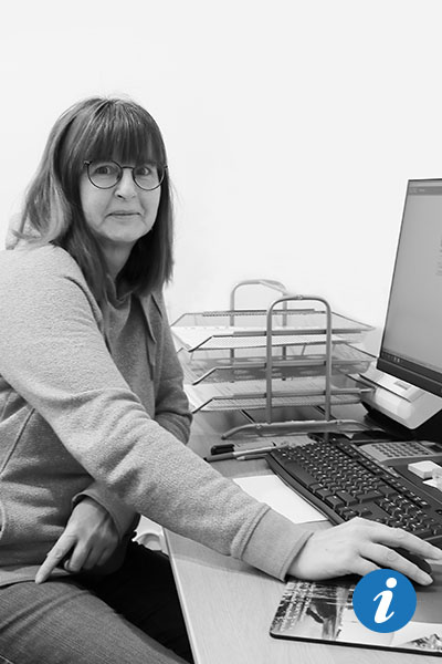 Eine Frau sitzt am Schreibtisch im Office der Physiotherapiepraxis EBENE2 und hält die Maus mit der rechten Hand, während sie in die Kamera guckt.