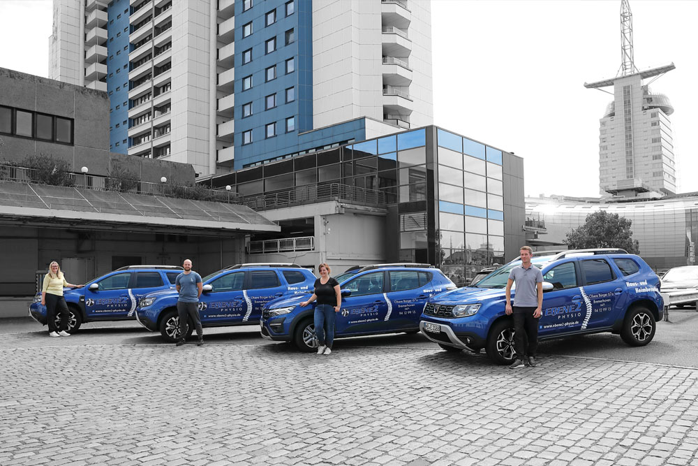 Hier sieht man vier Fahrzeuge der EBENE2 Physio vor dem Columbus Center in Bremerhaven. Die Physiotherapeuten posieren davor. Im Hintergrund sieht man das Sail City Hotel und das Klimahaus.