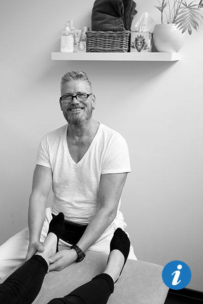 Ein Physiotherapeut setzt am Ende eine Behandlungsliege. Vor ihm liegt eine Patientin, von der man nur die Beine sehen kann. Der Physiotherapeut hat seine Hände am linken Fuß der Frau.